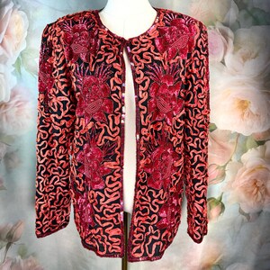 May include: A red and black sequined jacket with a floral design. The jacket is open in the front and has long sleeves. The floral design is made up of red sequins and black fabric. The jacket is displayed on a mannequin.