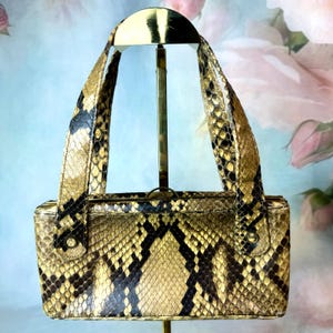 Puede incluir: Un bolso rectangular con un estampado de piel de serpiente en tonos beige, marrón y negro. El bolso tiene dos asas y un cierre dorado. El bolso se exhibe sobre un soporte dorado sobre un fondo floral suave.
