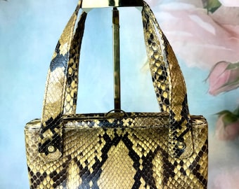 Könnte beinhalten: Eine rechteckige Handtasche mit Schlangenmuster in den Farbtönen Beige, Braun und Schwarz. Die Tasche hat zwei Henkel und einen goldfarbenen Verschluss. Die Tasche wird auf einem goldfarbenen Ständer vor einem weichen, floralen Hintergrund präsentiert.