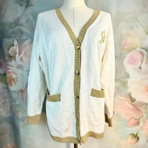 May include: Cream-colored cardigan with gold trim and buttons. The long-sleeved sweater features gold trim along the edges, pockets, and cuffs. A small gold emblem is embroidered on the upper left side. The cardigan is displayed on a mannequin.