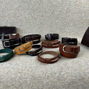 May include: A collection of ten leather belts in various colors, including brown, black, green, and yellow. The belts are arranged in a semi-circle on a light gray surface. Each belt has a silver buckle.