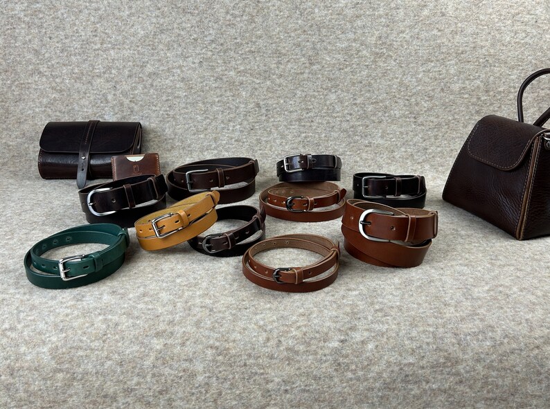 May include: A collection of ten leather belts in various colors, including brown, black, green, and yellow. The belts are arranged in a semi-circle on a light gray surface. Each belt has a silver buckle.