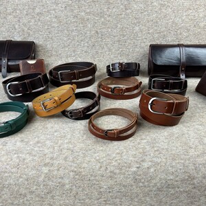 May include: A collection of leather belts in various colors, including brown, tan, green, and black. The belts are arranged in a circle on a light brown surface.