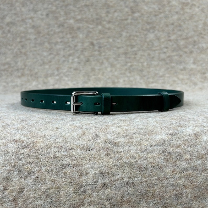 May include: A dark green leather belt with a silver buckle. The belt has multiple holes for adjusting the fit.