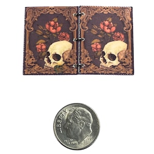 Puede incluir: Un pequeño diario de cuero marrón con un diseño de calavera y rosas. El diario tiene una encuadernación de anillas y es del tamaño de una moneda de diez céntimos de euro.