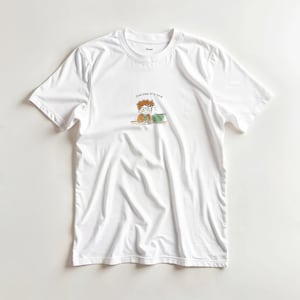 Peut inclure: T-shirt blanc à col rond avec une petite illustration d'une personne avec des lunettes travaillant sur un objet vert. Le texte "One step at a time" est au-dessus de l'illustration. Le t-shirt est en matière douce et légère.