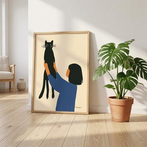 Könnte beinhalten: Gerahmter Kunstdruck mit einer stilisierten Illustration einer Person, die eine schwarze Katze hält. Die Person trägt ein blaues Hemd. Der Hintergrund ist hellbeige. Rechts vom Rahmen befindet sich eine Topfpflanze.