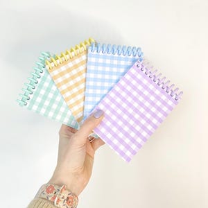 Peut inclure: Une main tenant cinq blocs-notes à spirales avec des motifs vichy pastel. Les couleurs sont vert menthe, jaune, bleu clair et lavande. La reliure spirale est blanche. Les blocs-notes sont disposés sur un fond blanc.