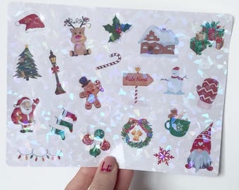 May include: A sheet of holographic Christmas stickers featuring festive designs. Includes Santa, a reindeer, a snowman, a gingerbread man, a Christmas tree, and a sign that says "Pole Nord". The stickers are in various colors and shapes.