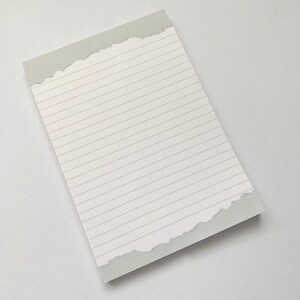 Può includere: Un blocco note a righe con un bordo strappato. La carta è bianca con linee grigie e il bordo superiore è grigio chiaro. Il blocco note è angolato su una superficie bianca.