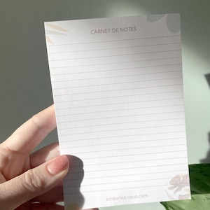 Peut inclure: Un bloc-notes blanc ligné avec le texte "CARNET DE NOTES" en haut et "emballez-vous.com" en bas. Le bloc-notes a un motif floral dans les coins. Le bloc-notes est tenu dans une main.