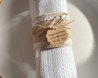 May include: A white napkin with blue stripes is tied with burlap ribbon and lace. A brown paper tag with the text "Virginie & Jérémy" and the date "09.09.2023" is attached to the ribbon.
