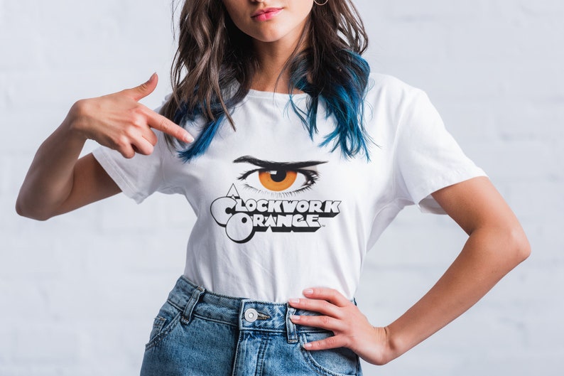 K&ouml;nnte beinhalten: Wei&szlig;es T-Shirt mit einer schwarz-wei&szlig;en Grafik eines Auges mit orangefarbener Iris und dem Text "A Clockwork Orange".