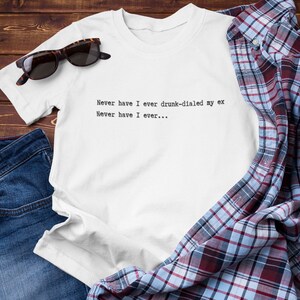 May include: White t-shirt with the text "Never have I ever drunk-dialed my ex. Never have I ever..." printed on the front. The t-shirt is laid out on a wooden surface with a pair of sunglasses and a blue denim shirt.
