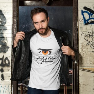 K&ouml;nnte beinhalten: Ein wei&szlig;es T-Shirt mit einem Aufdruck eines Auges und dem Text "A Clockwork Orange". Die Person, die das Shirt tr&auml;gt, steht in einem Durchgang mit Graffiti an den W&auml;nden.