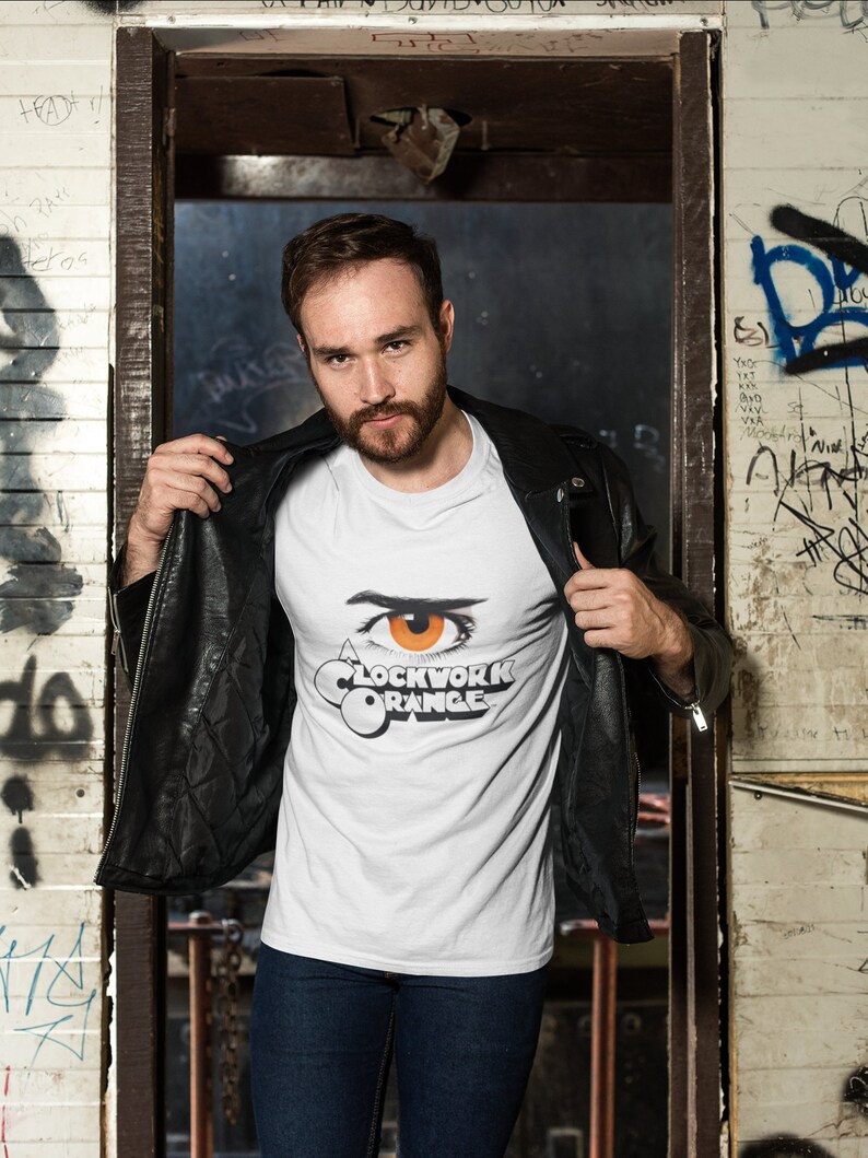 K&ouml;nnte beinhalten: Ein wei&szlig;es T-Shirt mit einem Aufdruck eines Auges und dem Text "A Clockwork Orange". Die Person, die das Shirt tr&auml;gt, steht in einem Durchgang mit Graffiti an den W&auml;nden.
