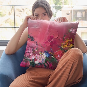 May include: A pink and red throw pillow with a floral design. The pillow has a textured, brushstroke-like pattern and features a variety of flowers in shades of pink, red, yellow, and green.