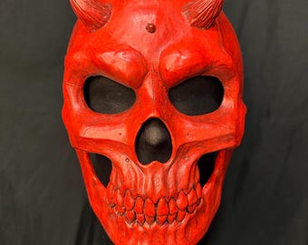 Puede incluir: Una máscara de calavera de color rojo brillante que presenta dos pequeños cuernos texturizados que sobresalen de la zona de la frente. La máscara tiene cuencas oculares profundas y huecas, y una hilera de dientes detallada en la mandíbula.