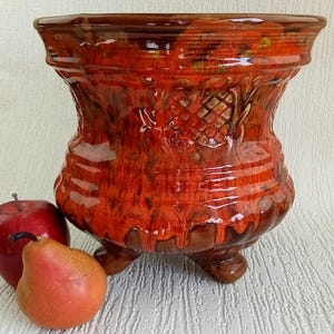 May include: A handcrafted, glazed ceramic planter in shades of red and brown, featuring a textured surface and a decorative embossed design. The planter has a wide, flared rim and rests on three small feet. An apple and a pear are next to the planter.