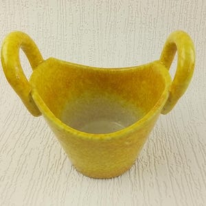 May include: A yellow ceramic planter with two handles. The planter has a speckled texture and a rounded shape.