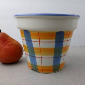May include: A ceramic flower pot with a white top and a colorful plaid pattern in orange, blue, and green. A pear is next to the pot.