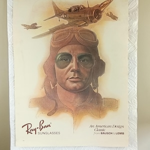 May include: Vintage Ray-Ban sunglasses advertisement featuring a pilot wearing aviator sunglasses and a leather helmet. The image includes a sepia-toned illustration of a plane and the text "An American Design Classic from BAUSCH & LOMB."