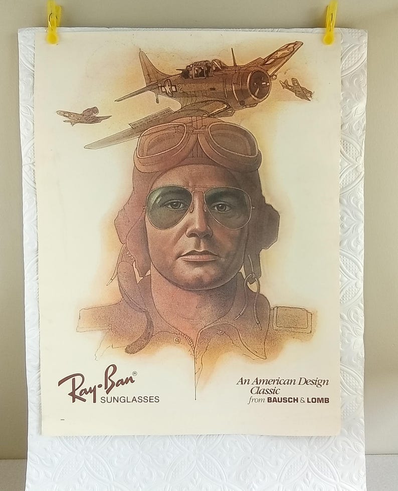 May include: Vintage Ray-Ban sunglasses advertisement featuring a pilot wearing aviator sunglasses and a leather helmet. The image includes a sepia-toned illustration of a plane and the text "An American Design Classic from BAUSCH & LOMB."