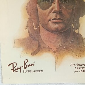 May include: An illustration of a person wearing aviator sunglasses. The Ray-Ban logo is in the lower left corner, with the words "SUNGLASSES" below it. The text "An American Classic from BAUSCH & LOMB" is in the lower right corner.