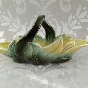 May include: A ceramic basket in a leaf design, featuring a handle and a two-tone color scheme. The basket has a light yellow interior and is surrounded by green and dark green leaves.