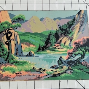May include: A vintage paint-by-number landscape scene featuring a serene lake surrounded by mountains and lush greenery. The artwork showcases a small house nestled among the trees, with a detailed depiction of the natural environment. The color palette includes shades of green, blue, and brown.
