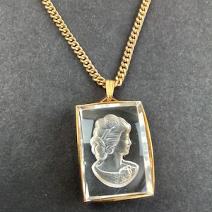 May include: A gold-toned necklace with a rectangular pendant. The pendant features a clear glass or crystal with a carved profile of a woman. The chain is a curb link style.