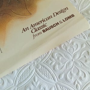 May include: A close-up of a cream-colored paper with the text "An American Design Classic from BAUSCH & LOMB" in brown. The paper is on a white, textured surface with a floral pattern.