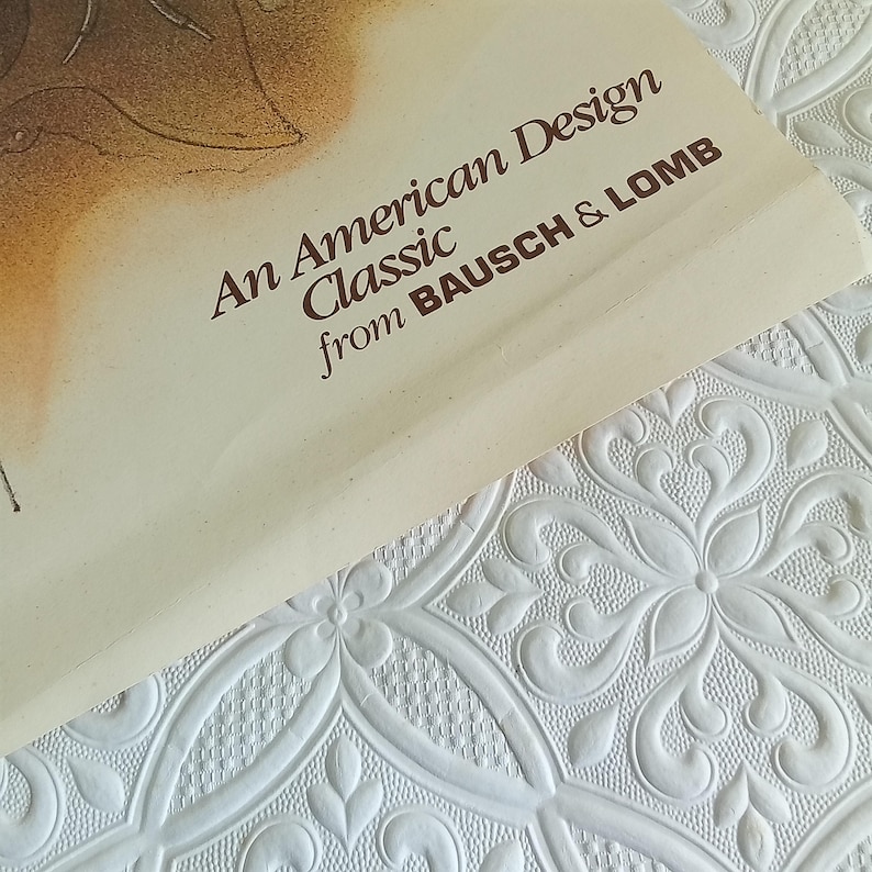 May include: A close-up of a cream-colored paper with the text "An American Design Classic from BAUSCH & LOMB" in brown. The paper is on a white, textured surface with a floral pattern.