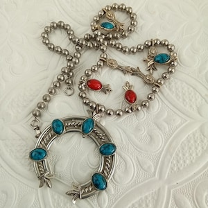 May include: Silver-toned necklace and bracelet set with turquoise and red stone accents. The necklace features a horseshoe pendant with turquoise stones. The bracelet and necklace have a beaded design with decorative gold-toned elements.