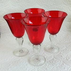 May include: A set of four wine glasses featuring deep red, translucent bowls with a subtle bubbled texture. Each glass is supported by a clear, smooth glass stem and a circular base, creating a uniform look across the entire collection of glassware.