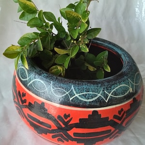 May include: A round, hand-painted ceramic planter with a wide opening. The planter features a red band with black geometric designs, a blue band with white accents, and is filled with green leafy branches.