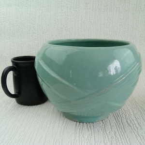May include: A teal ceramic bowl with a rounded shape and a decorative, crisscrossing design. A black mug with a handle is next to the bowl. The bowl has a glossy finish and sits on a white surface.