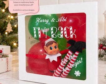 May include: A white rectangular box with a clear window displaying a toy elf with a red hat and striped legs, surrounded by red and green balloons. The box features the text Harry & Abi, I'M BACK, and Let the fun begin! with a snowflake icon.
