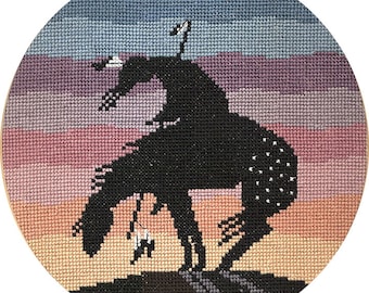 May include: Cross-stitch embroidery of a rodeo cowboy on a bucking bronco. The black silhouette of the rider and horse is set against a sunset of blue, purple, pink, and orange hues. The artwork is framed in a round wooden hoop.