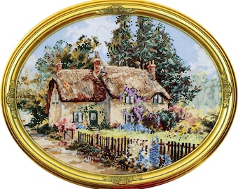 May include: A cross-stitch embroidery of a quaint cottage with a thatched roof, surrounded by lush greenery and a picket fence. The cottage is painted in a soft white with a green door and windows. The scene is framed in a gold oval frame.
