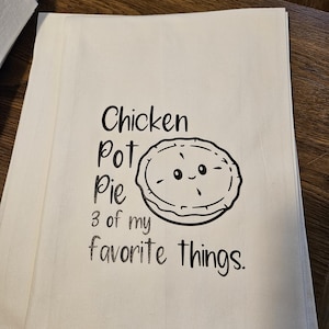 May include: A white kitchen towel with black text that reads "Chicken Pot Pie 3 of my favorite things." A black and white cartoon pie with a smiling face is next to the text.