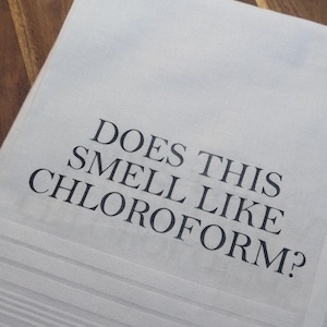 May include: White cotton tea towel with black text that reads "Does this smell like chloroform?"