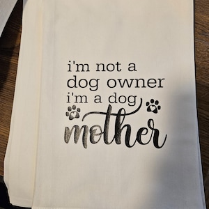 May include: A stack of off-white kitchen towels with the text "I'm not a dog owner I'm a dog mother" printed in black. The phrase includes paw print graphics. The towels are folded and stacked on a wooden surface.