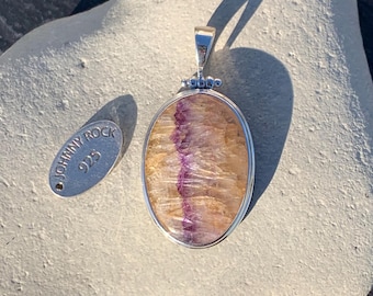 May include: Oval pendant with a silver setting, featuring a polished stone with a central purple stripe and golden hues. A small silver tag with the text "JOHNNY ROCK 925" is also present. The pendant is a piece of jewelry.