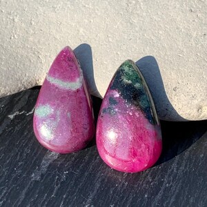 Op de afbeelding: Twee traanvormige roze en groene edelsteen cabochons. De cabochons hebben een glad, gepolijst oppervlak en zijn geplaatst tegen een donkere achtergrond.