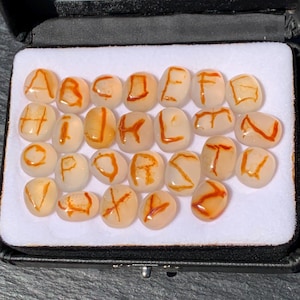 Op de afbeelding: Een set van zesentwintig gladde, doorschijnende, gebroken witte ovale stenen in een zwarte displaydoos met een witte schuimrubberen inzet. Elke steen is voorzien van een handgeschilderde oranje letter van het alfabet van A tot Z.