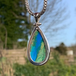 Puede incluir: Un colgante en forma de l&aacute;grima con una piedra preciosa azul y verde engastada en un marco de plata. El colgante est&aacute; colgado de una cadena de plata.