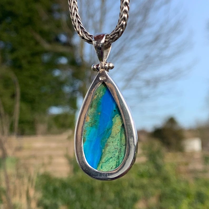 Puede incluir: Un colgante en forma de l&aacute;grima con una piedra preciosa azul y verde engastada en un marco de plata. El colgante est&aacute; colgado de una cadena de plata.