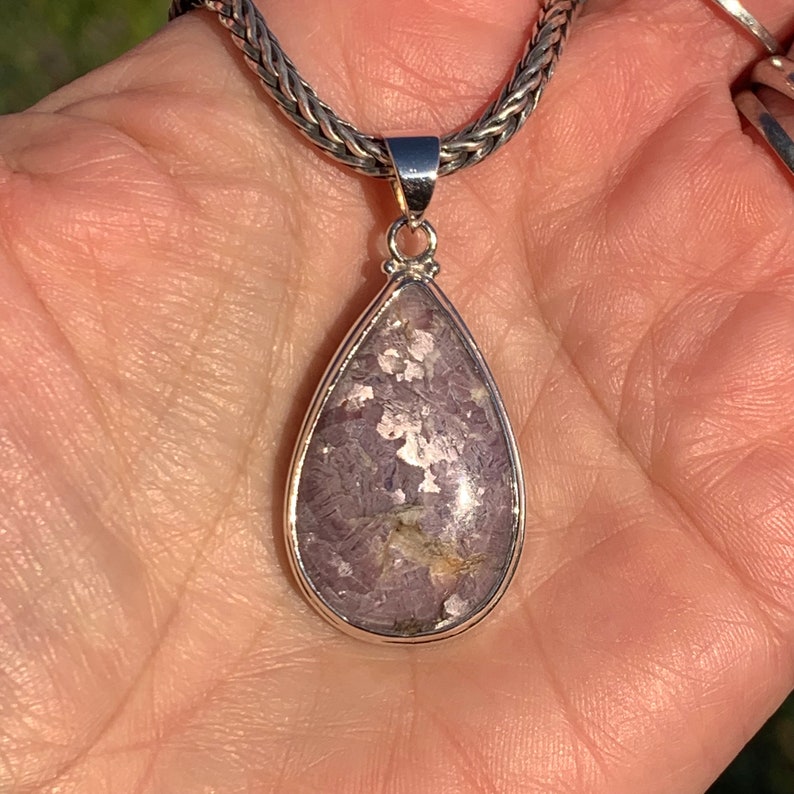 K&ouml;nnte beinhalten: Ein tropfenf&ouml;rmiger Anh&auml;nger mit einer silbernen Kette. Der Anh&auml;nger zeigt einen violetten und silbernen Edelstein mit einem glitzernden Finish.