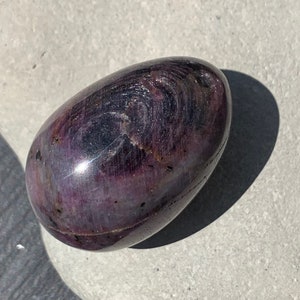 Könnte beinhalten: Ein polierter, oval geformter Stein mit tiefvioletter Farbe und wirbelnden Mustern. Der Stein hat eine glänzende Oberfläche und liegt auf einer hellgrauen Oberfläche. Die Farbtöne reichen von Dunkelviolett bis zu helleren Schattierungen.